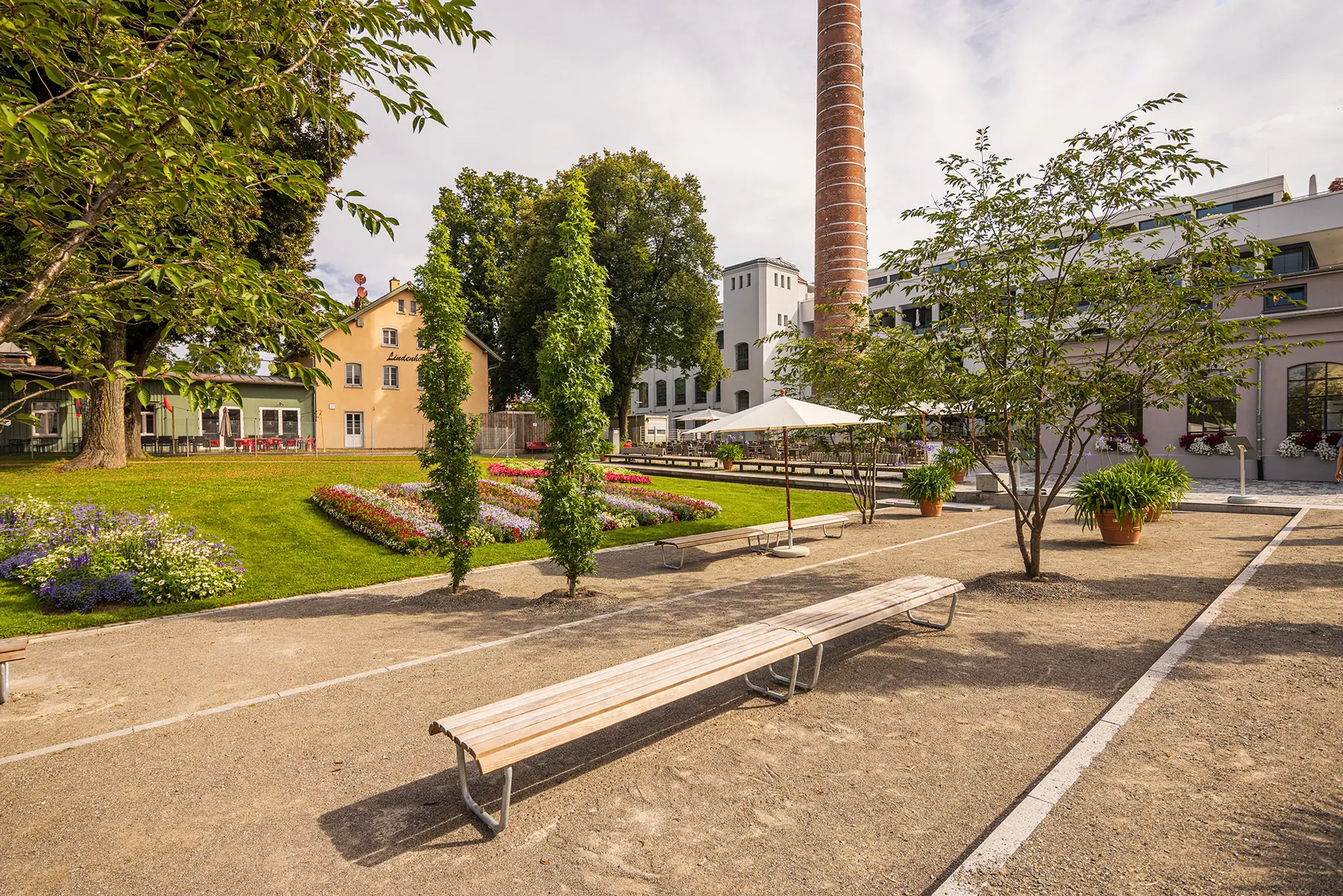 Grüner Platz mit Sitzbänken vor der historischen Spinnerei und ihrem Backsteinschornstein